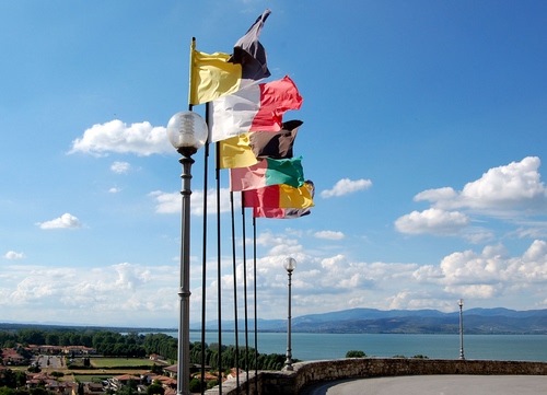 Immagine con bandiere, cielo, nuvole, lampioni, colori, case, aste, acqua, mare, alberi, verde, azzurro, panorama, vento, rosso, lago